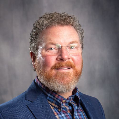 A man with a beard and glasses is wearing a blue suit and plaid shirt.