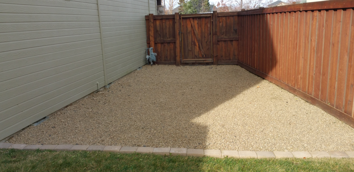 There is a gravel area in the backyard next to a wooden fence.