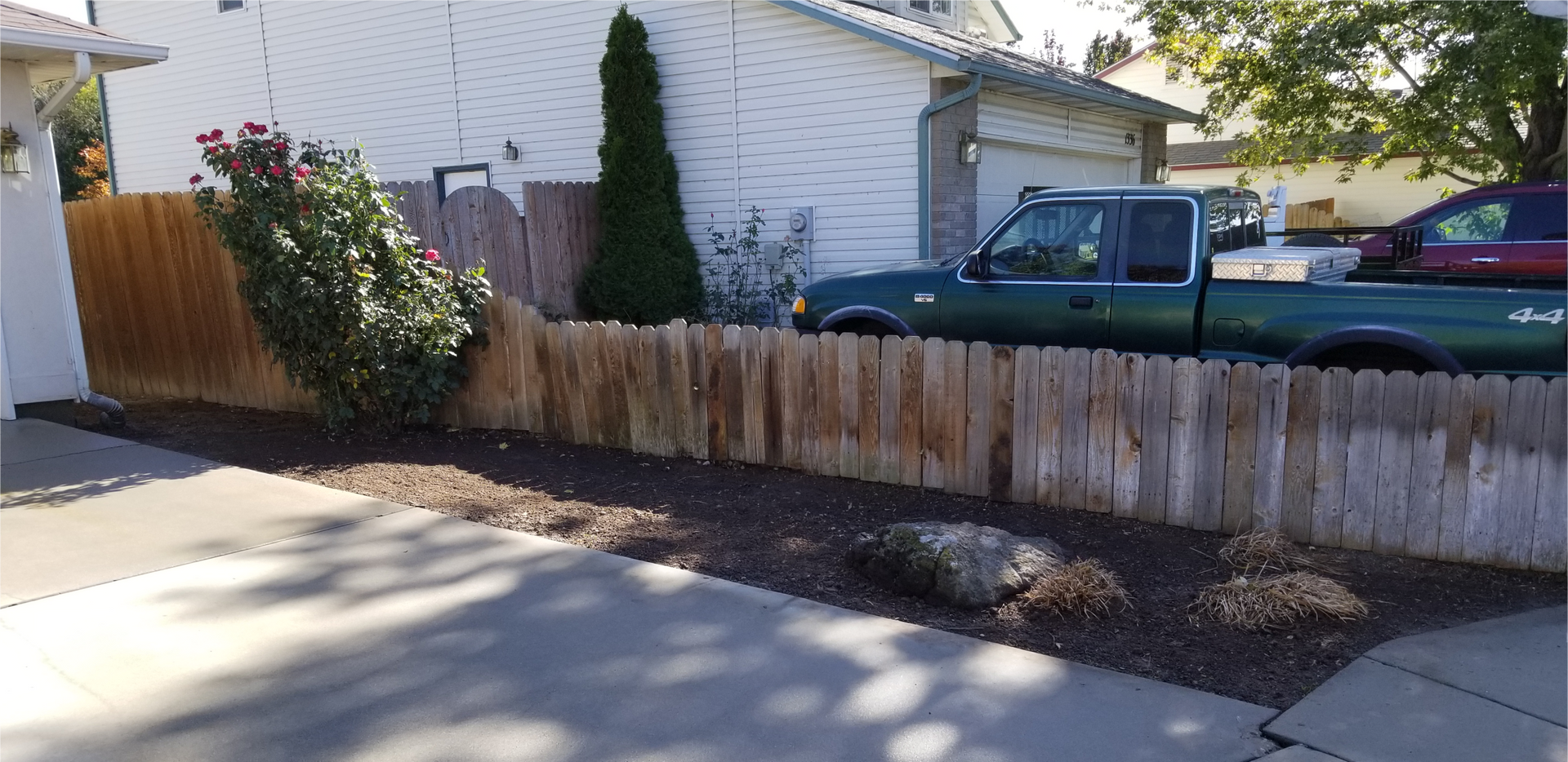 A wooden fence lines a driveway, behind it a green truck and house are visible.