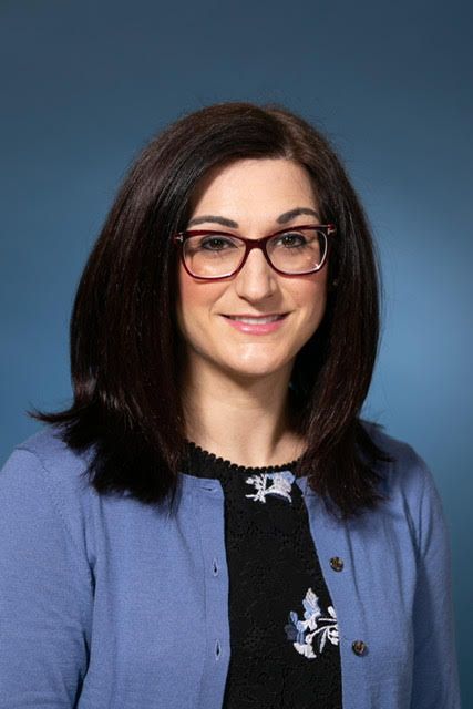 A woman wearing glasses and a blue cardigan is smiling for the camera.