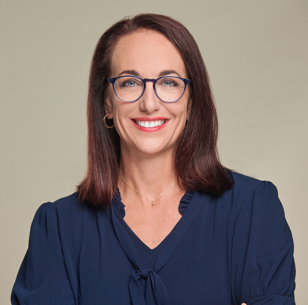 A woman wearing glasses and a blue shirt is smiling.