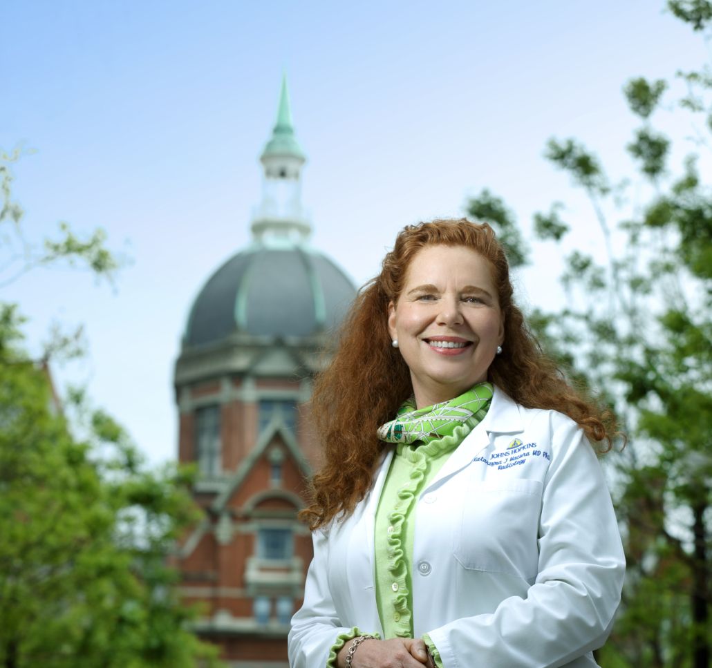 A woman in a white coat and green scarf is smiling in front of a dome shaped building
