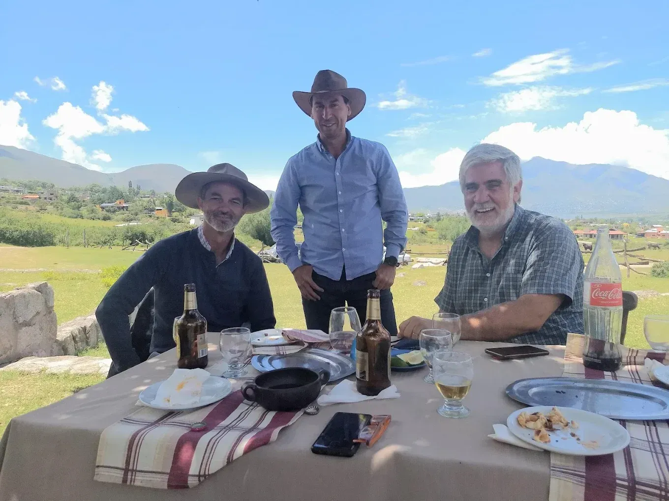 Tres hombres en una mesa al aire libre; uno con sombrero de vaquero, montañas al fondo, día soleado.