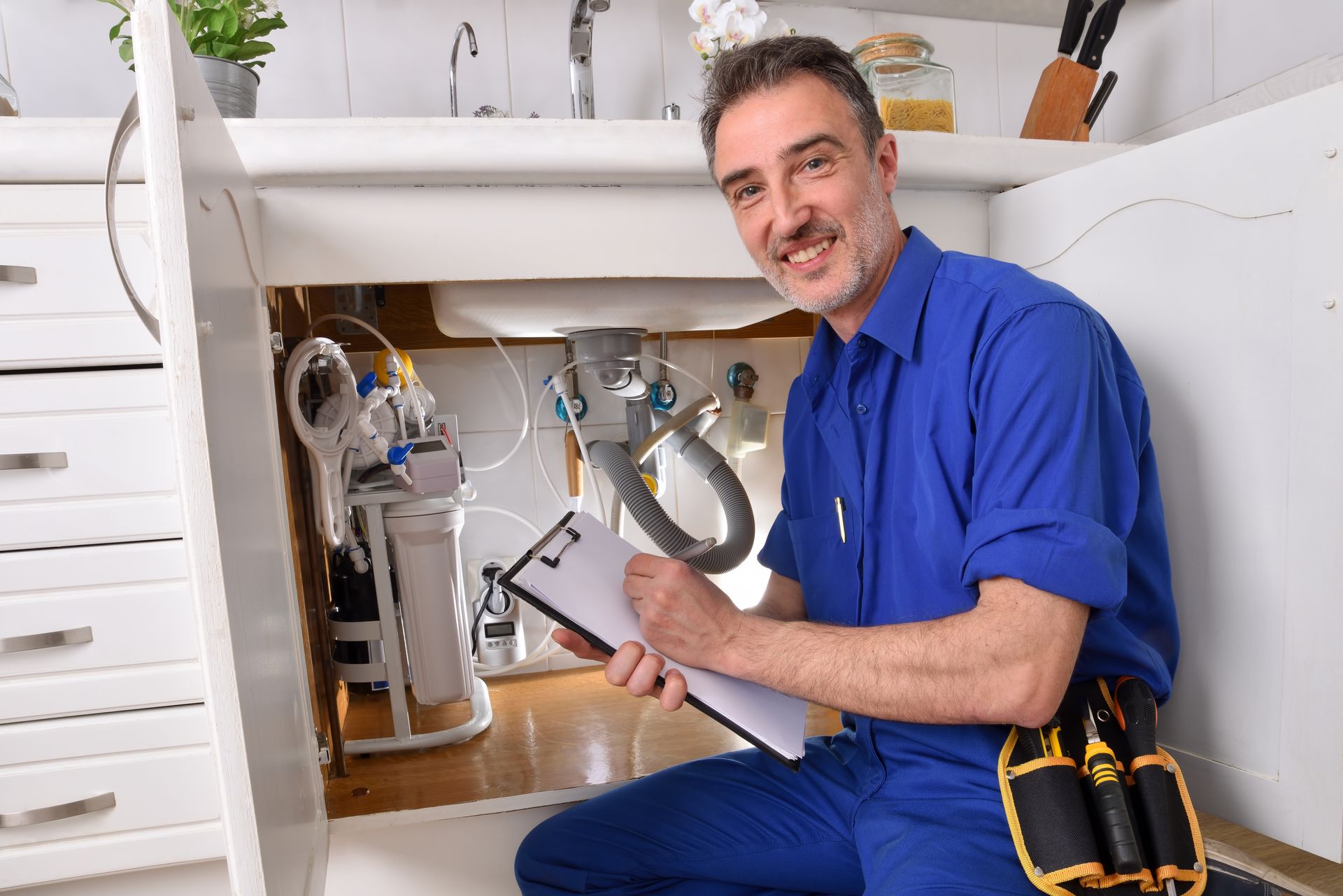 Plumber in blue uniform, kneeling under kitchen sink, holding clipboard, smiling.