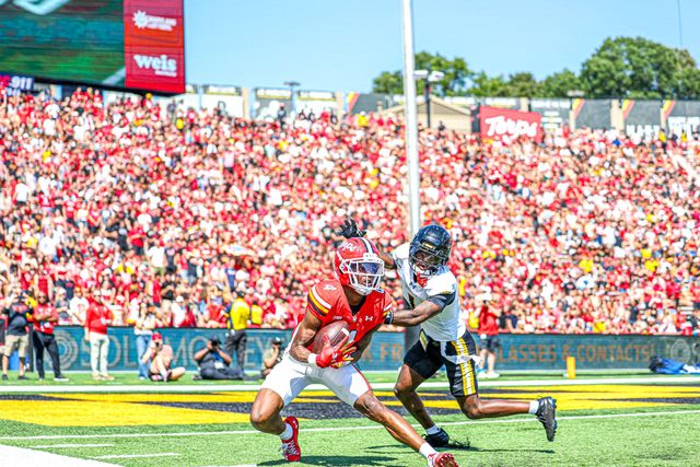 A college football player in a red uniform carries the ball past a defender in white during a game in a crowded stadium.
