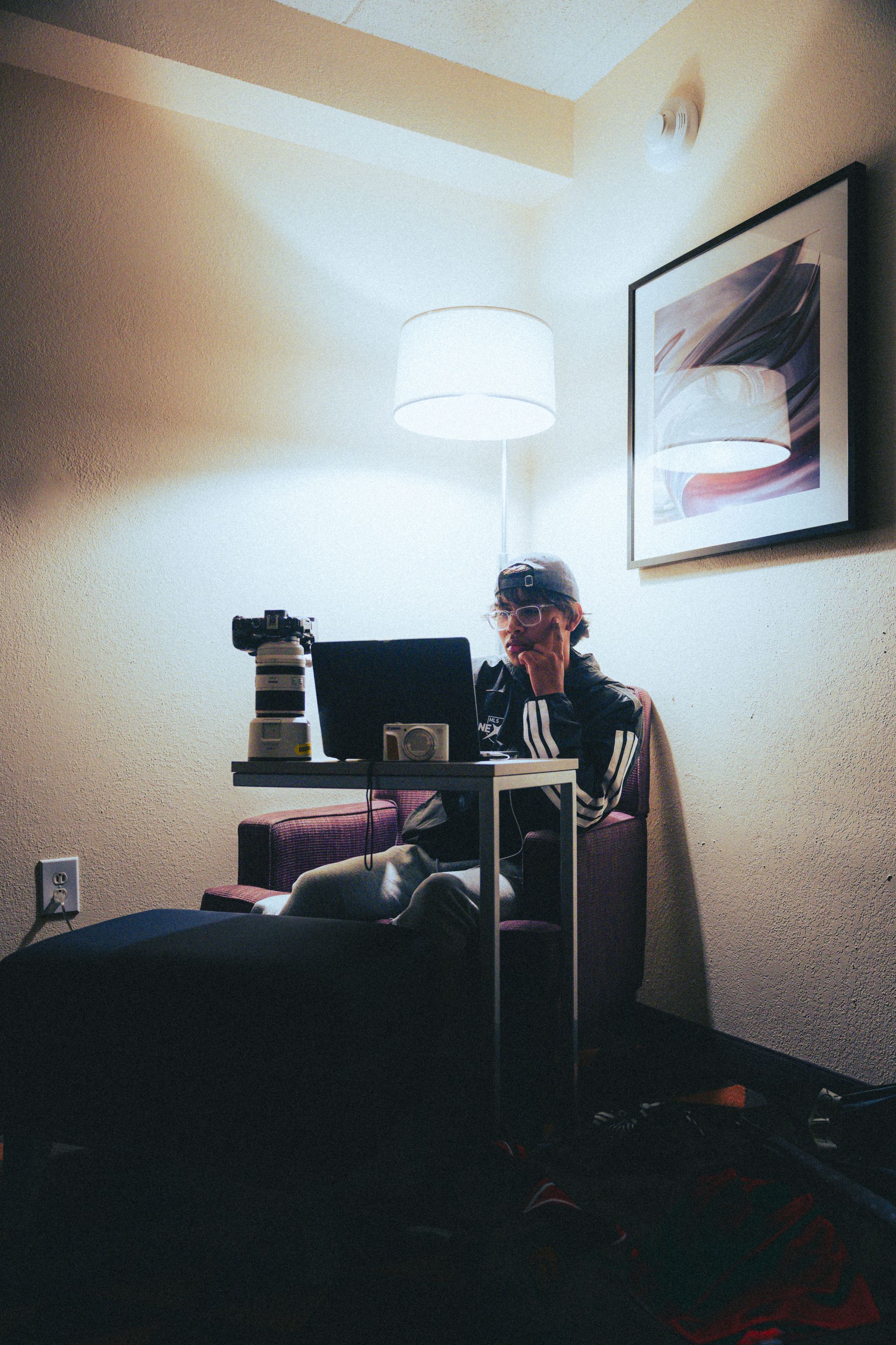 Person seated at a desk in a dim room, with a lamp, monitor, and framed wall art.