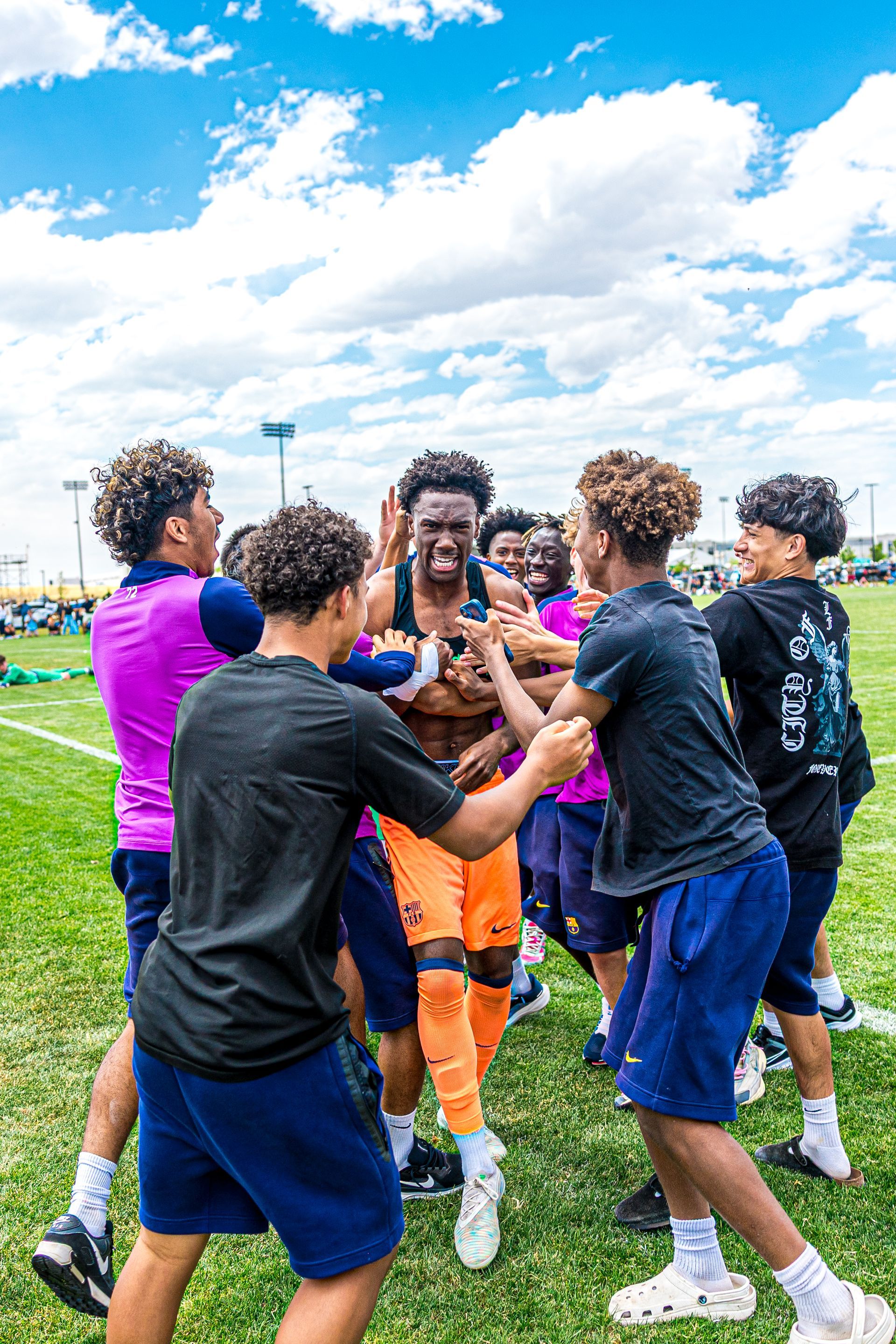 Soccer players huddle and celebrate on a grassy field under a bright blue sky.
