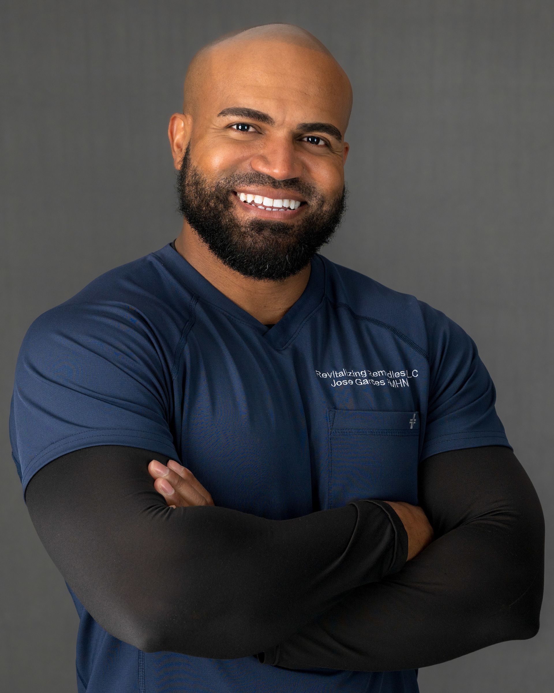 Bald man with beard, arms crossed, smiling. Wearing dark blue top with black sleeves. Against gray backdrop.
