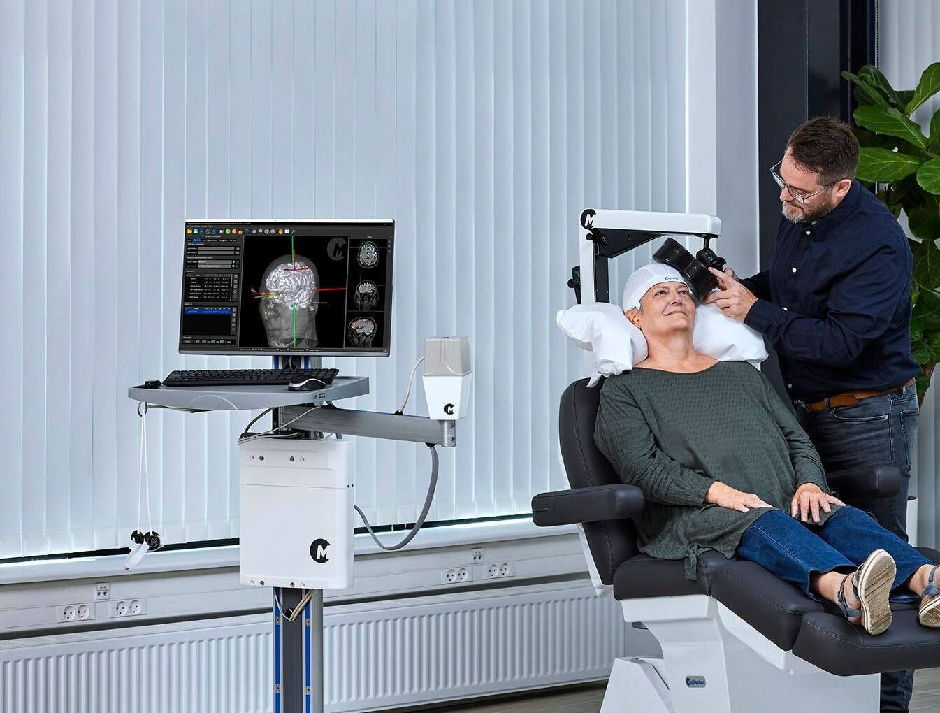 A technician adjusts a cap on a person seated in a chair next to a brain-scanning device. The setting is indoors.