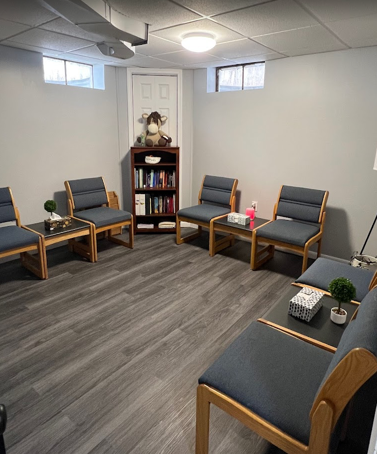 Therapy room with gray chairs arranged in a circle, small tables, bookcase, and gray flooring.