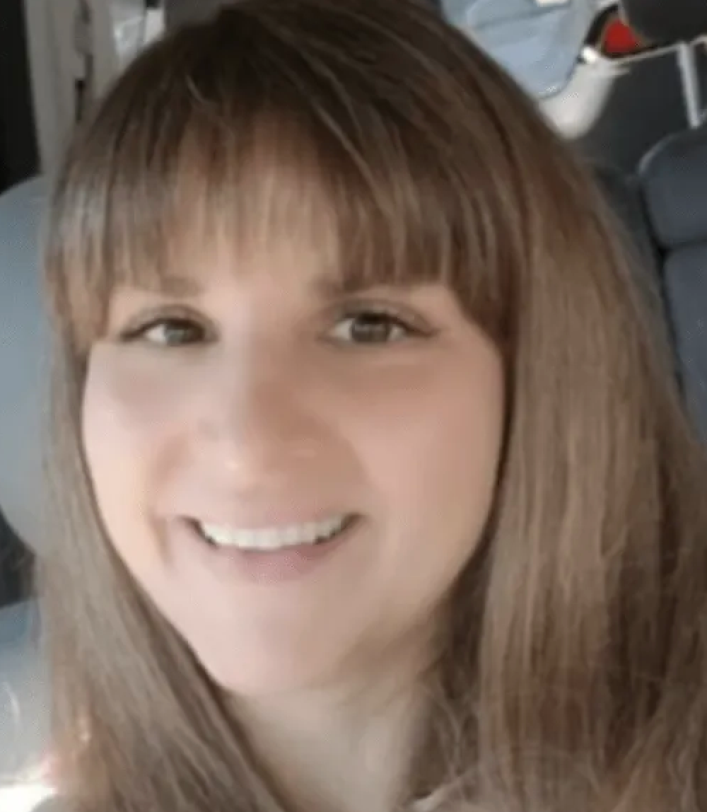 Woman with long brown hair and bangs, smiling in a car.