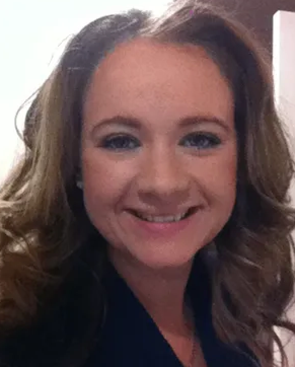 Woman with light brown wavy hair smiles, wearing a dark blazer; looking directly at the viewer.