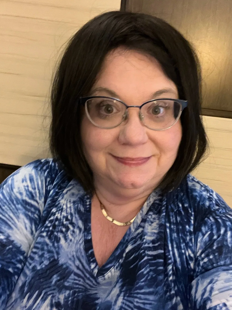 Woman with glasses, dark hair, and blue tie-dye shirt smiles at the camera.
