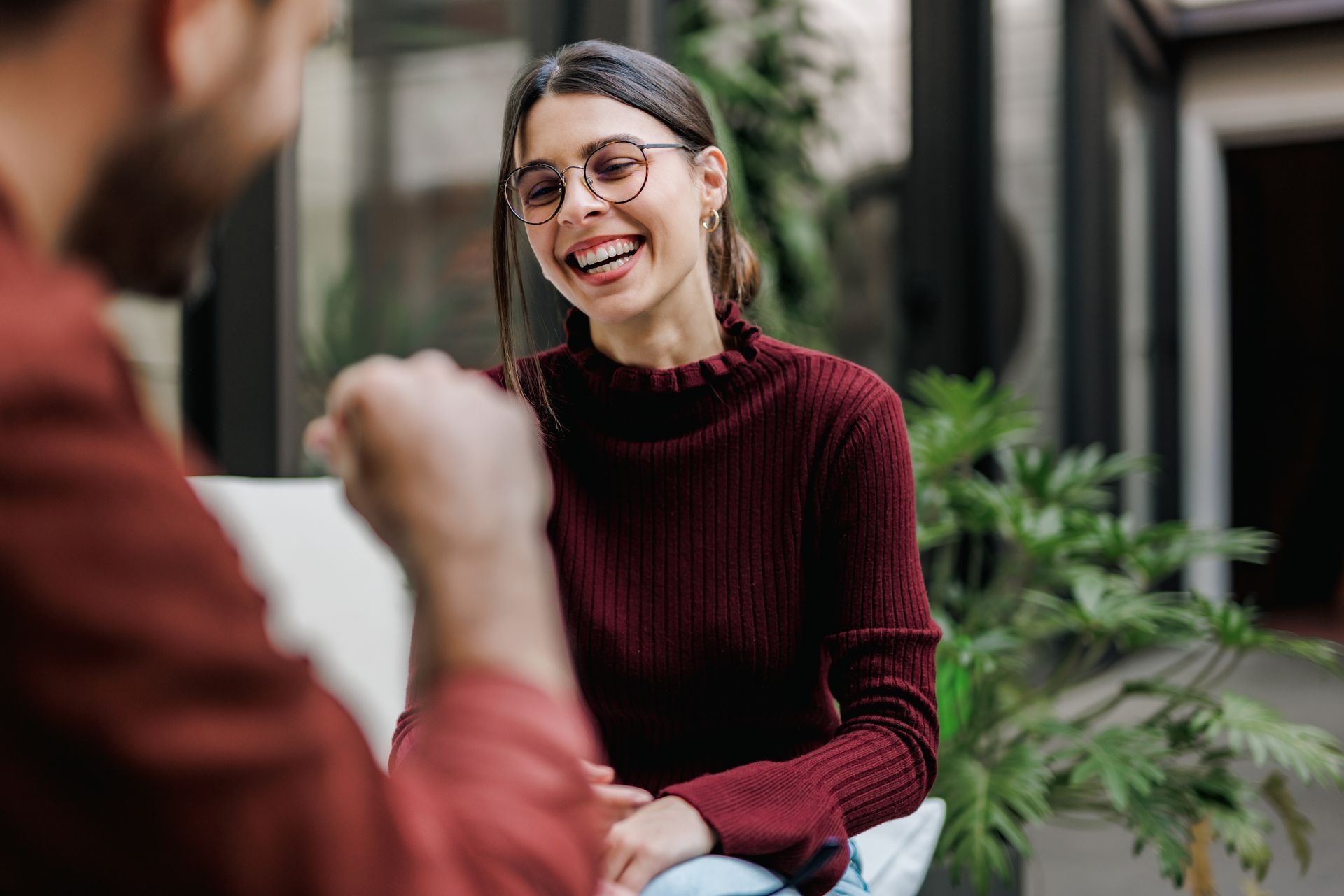 Woman in maroon sweater laughs while talking to a person out of focus; setting has plants and a window.