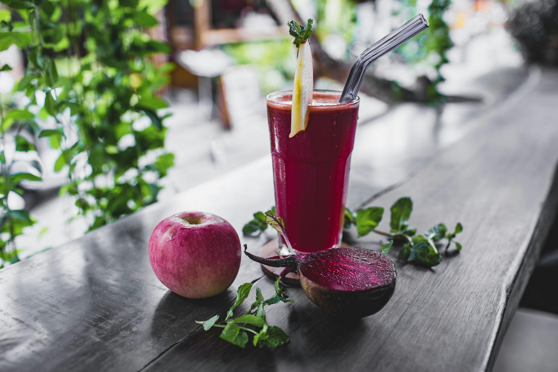 Glass of red juice with straw, apple, and halved beet on wooden counter.
