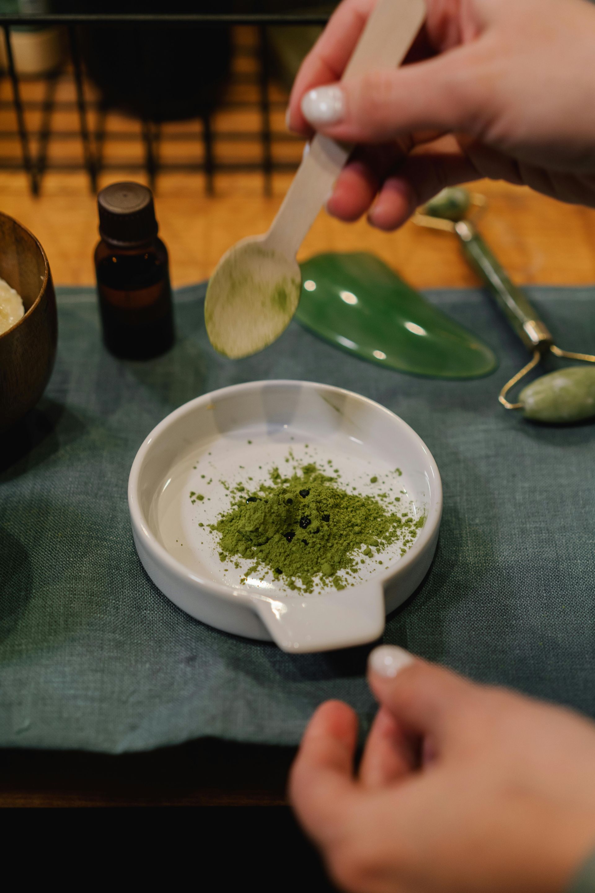 Person adding green powder to a small white bowl, skincare preparation. Green jade roller, oil bottle on blue cloth.