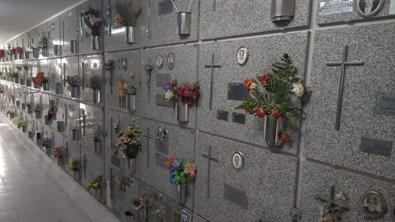 Vista interior de un muro de mausoleo con numerosos nichos individuales que albergan urnas y flores. Piedra gris con cruces.