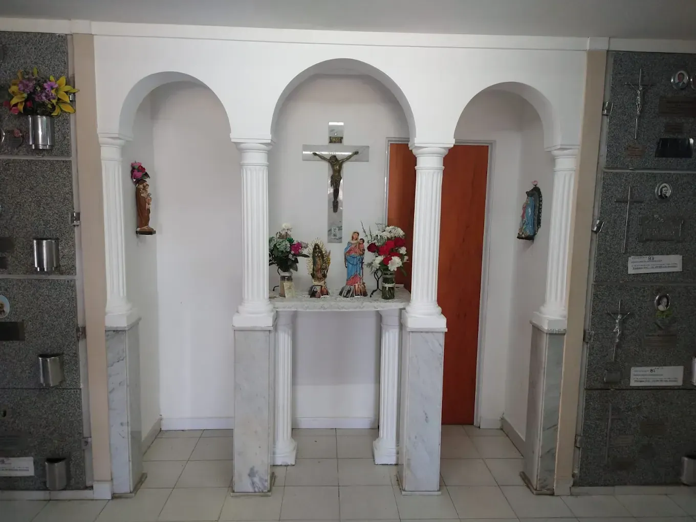 Una alcoba en un mausoleo que alberga estatuas religiosas y un crucifijo, flanqueada por criptas de granito gris.