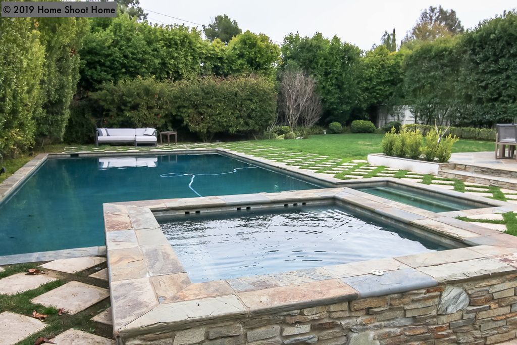 Backyard pool and spa surrounded by stone and lawn, with trees in the background.