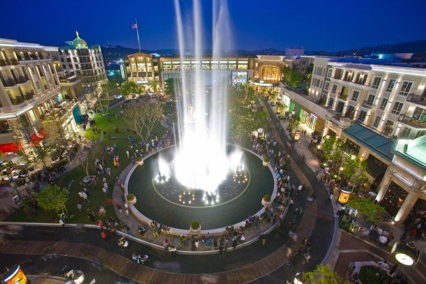 Night view of a mall with a large water fountain surrounded by people and shops, with bright lights.