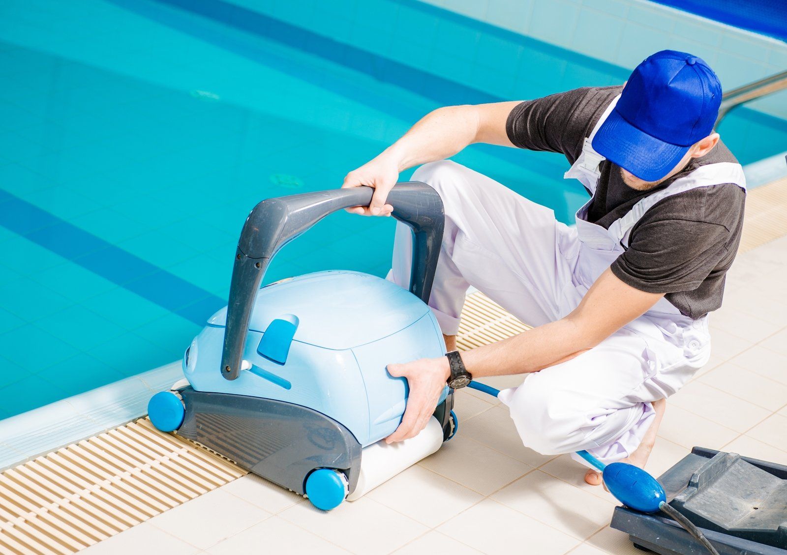 Man in blue cap and white overalls, cleaning a pool with an automated cleaner.