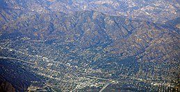 Aerial view of a city nestled between brown mountains. Roads, buildings, and trees are visible in the valley.