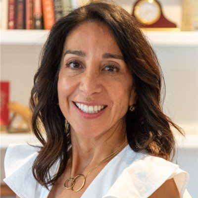 Woman with dark hair smiles, wearing a white top and gold necklace, bookshelves in the background.