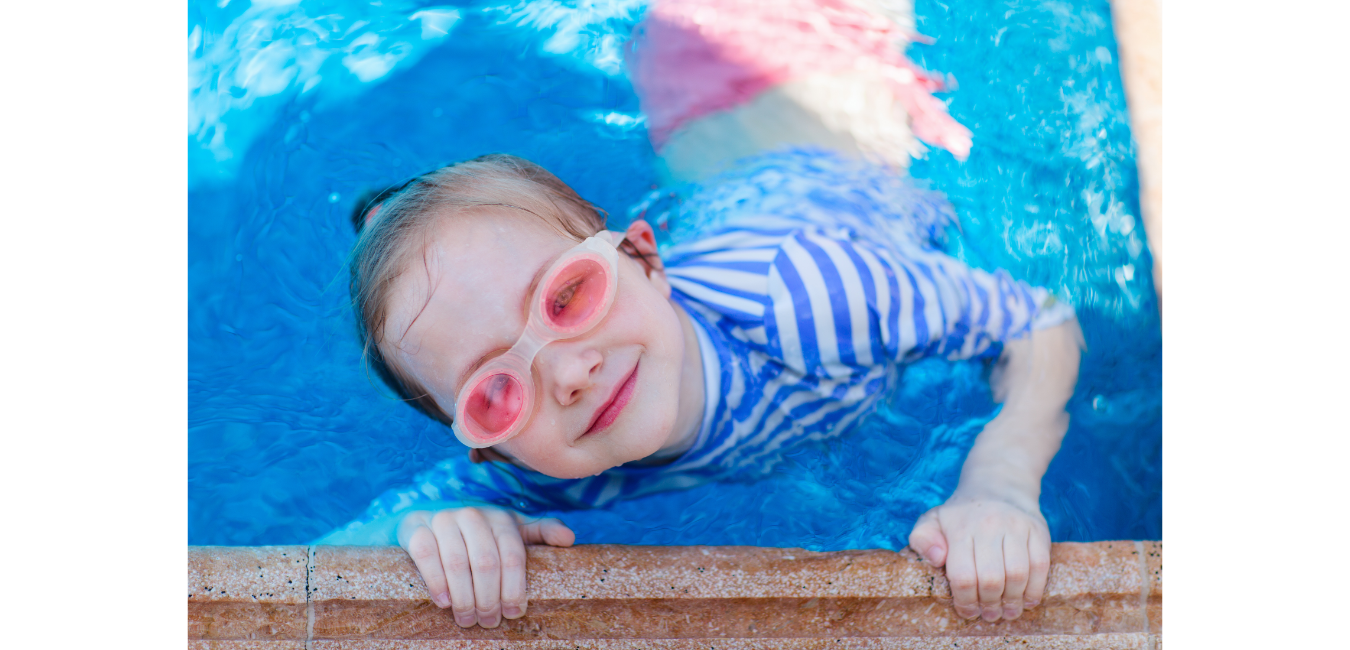 Child wearing pink goggles and striped shirt smiles in a blue pool.