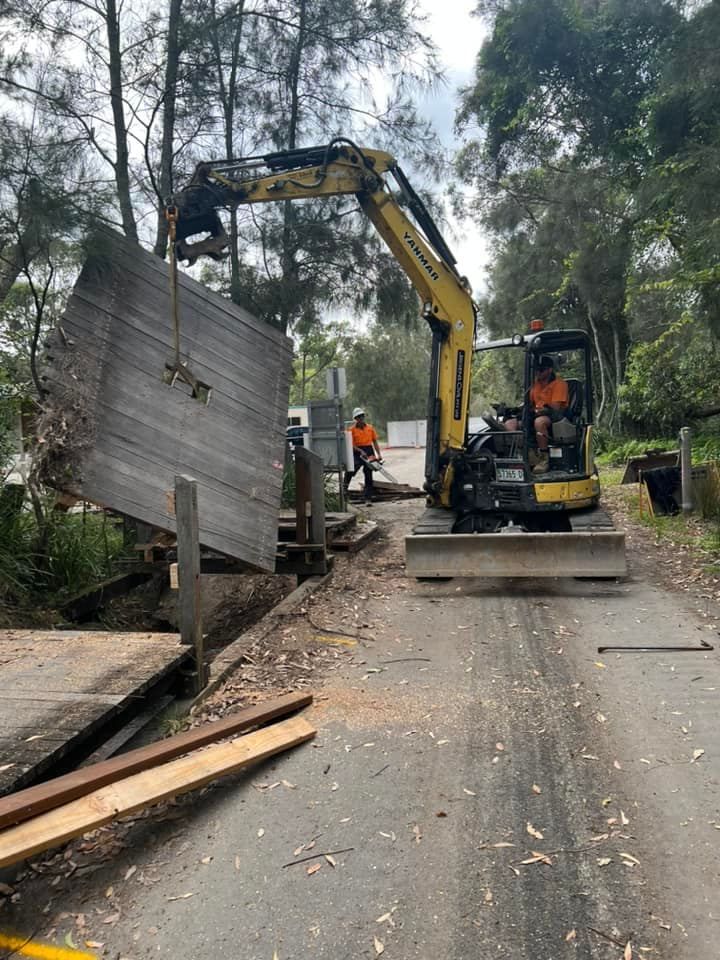 Yellow Excavator Lifting a Wooden Barrier on A Road, with A Worker Nearby — ALI Civil Pty Ltd in Sussex Inlet, NSW