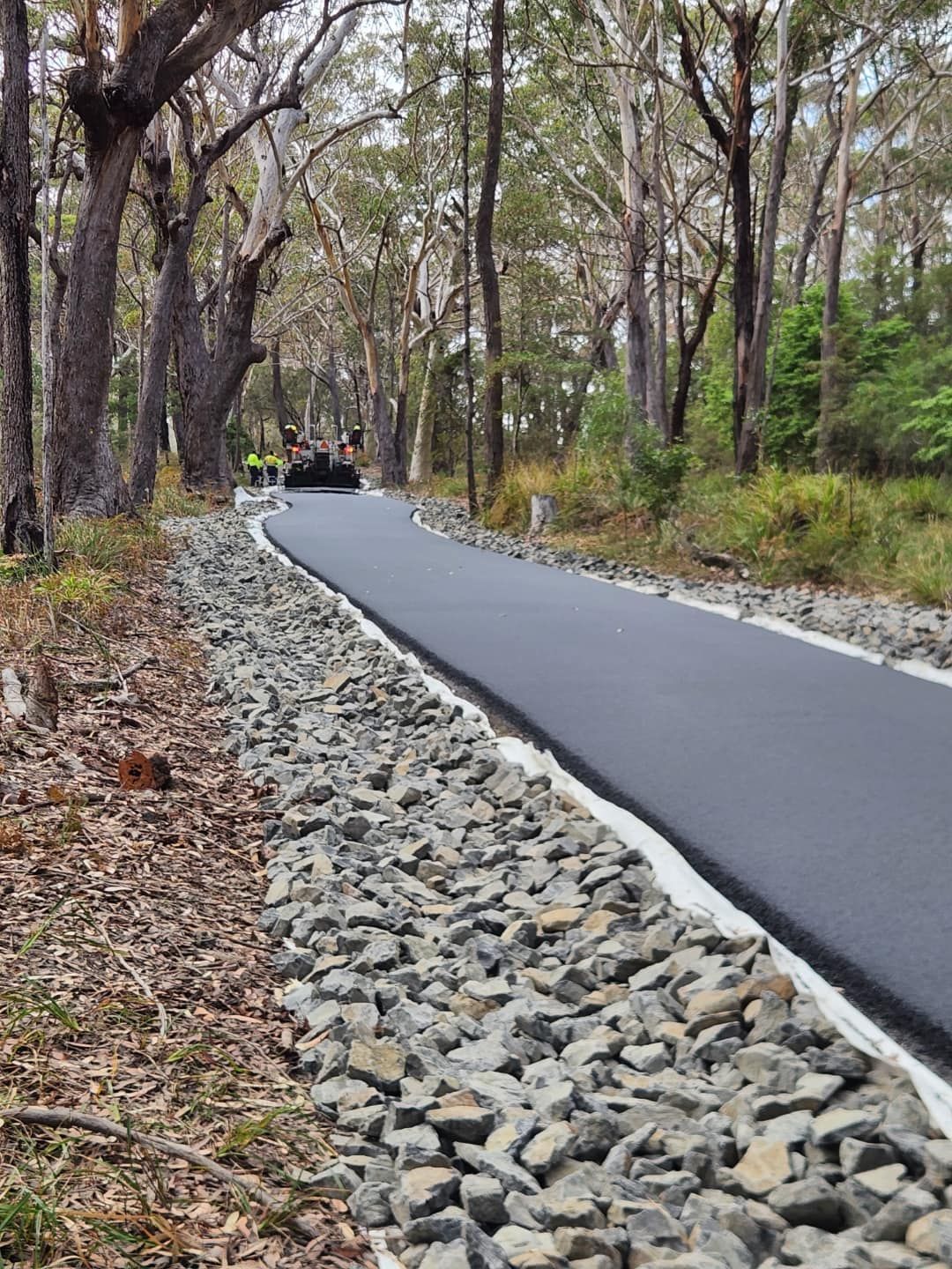 Asphalt path winding through a forest, edged with gravel and bordered by trees. A machine works on the path. — ALI Civil Pty Ltd in South Nowra, NSW