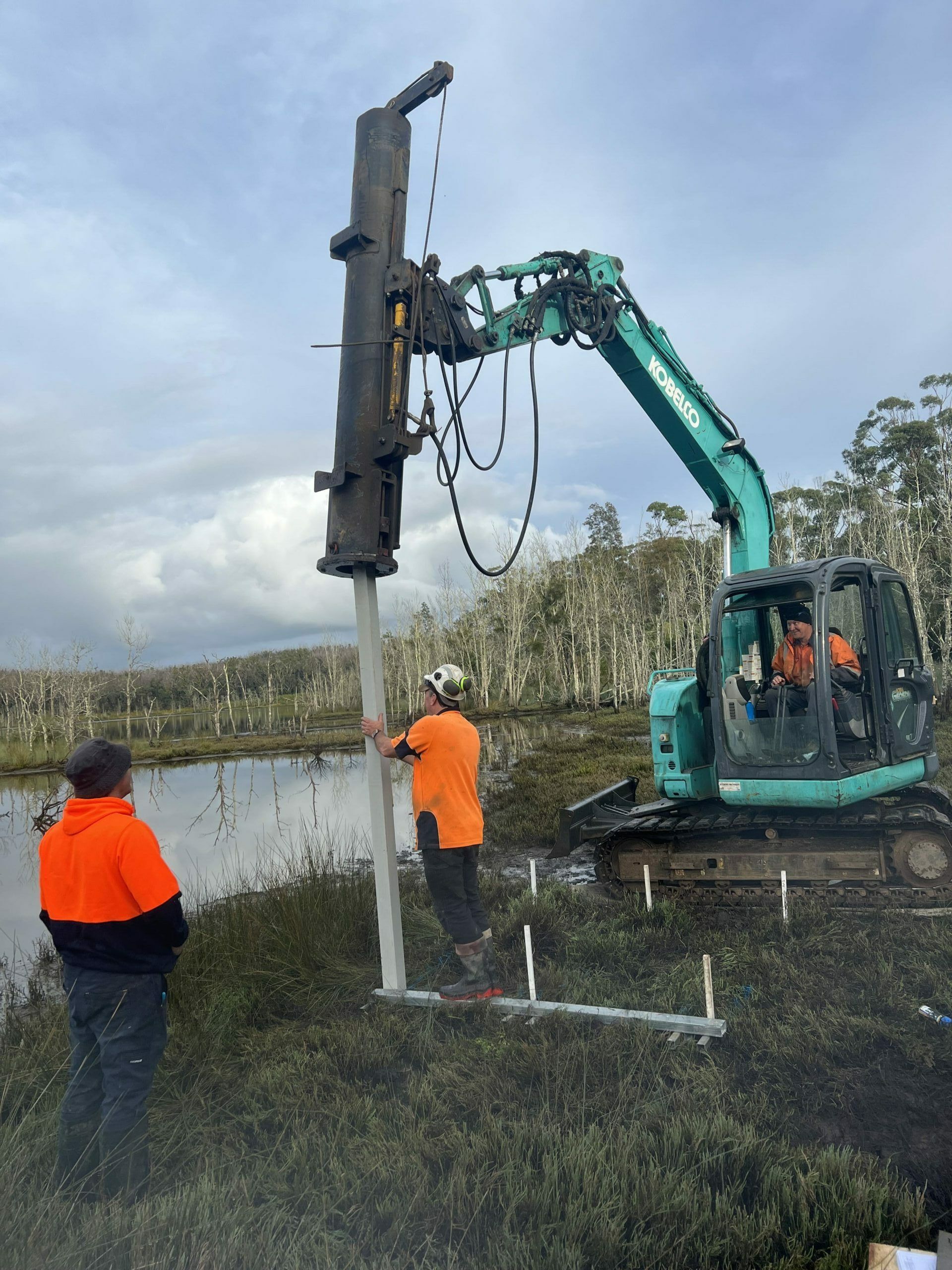Workers using an excavator to install a white pole in a wetland area. One worker helps guide the pole. — ALI Civil Pty Ltd in South Nowra, NSW