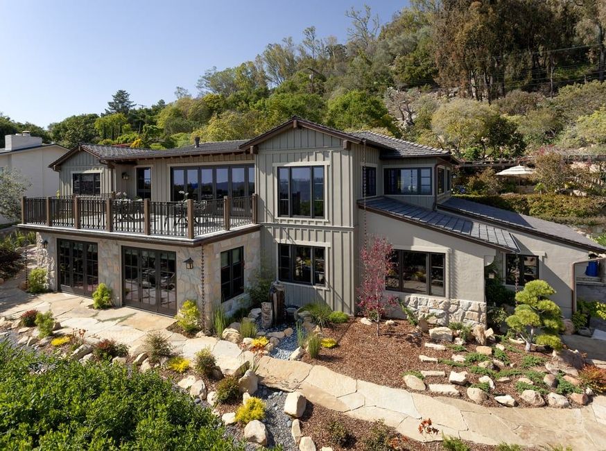 A multi-story home with vertical siding and stone accents, featuring a large deck and terraced landscaping in a hilly area.