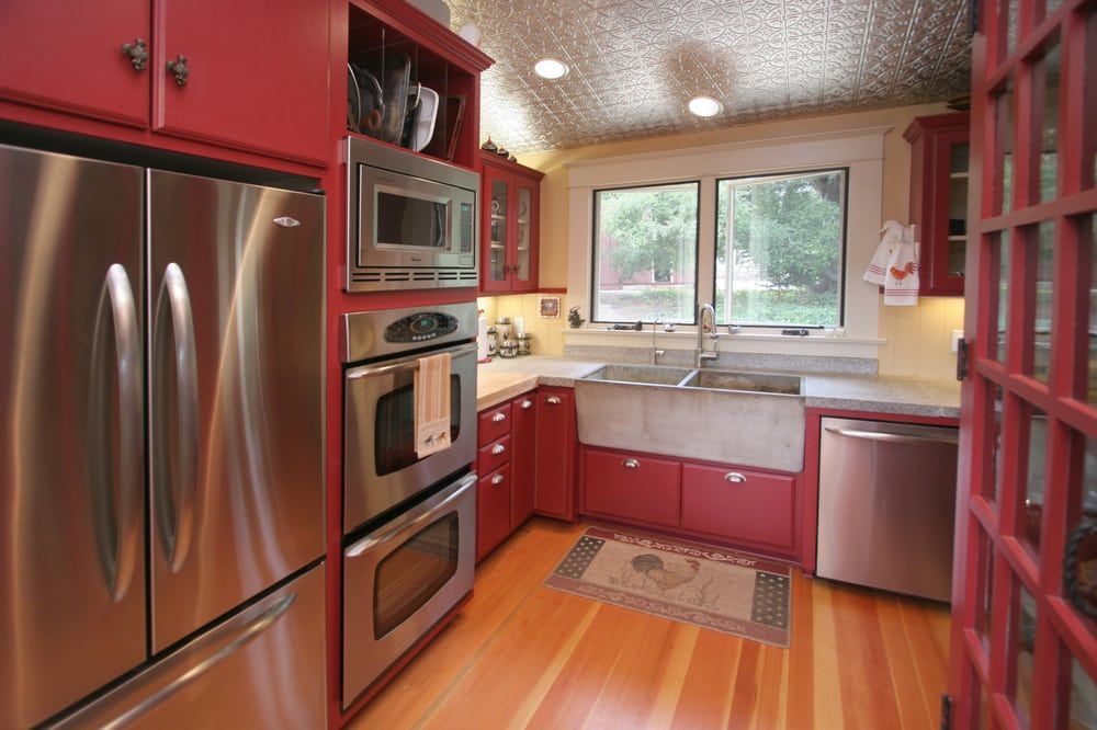 A cozy, narrow kitchen featuring bright red cabinets, stainless steel appliances, a farmhouse sink, and a tin ceiling.