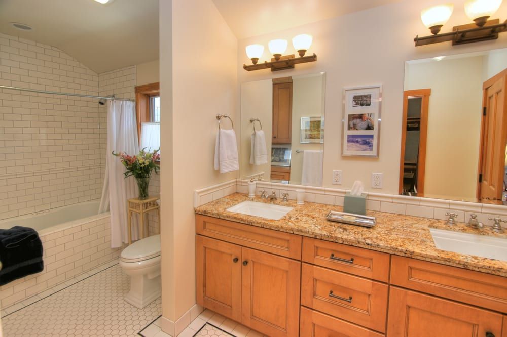 A bright bathroom featuring a double vanity with a granite countertop, wood cabinets, and a white subway-tile shower.
