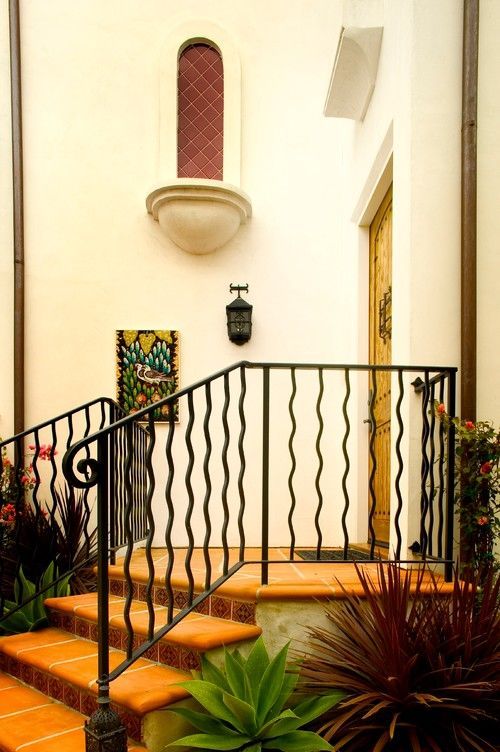 Terracotta steps with black wrought-iron railing leading to a white stucco entryway with plants and a stained-glass window