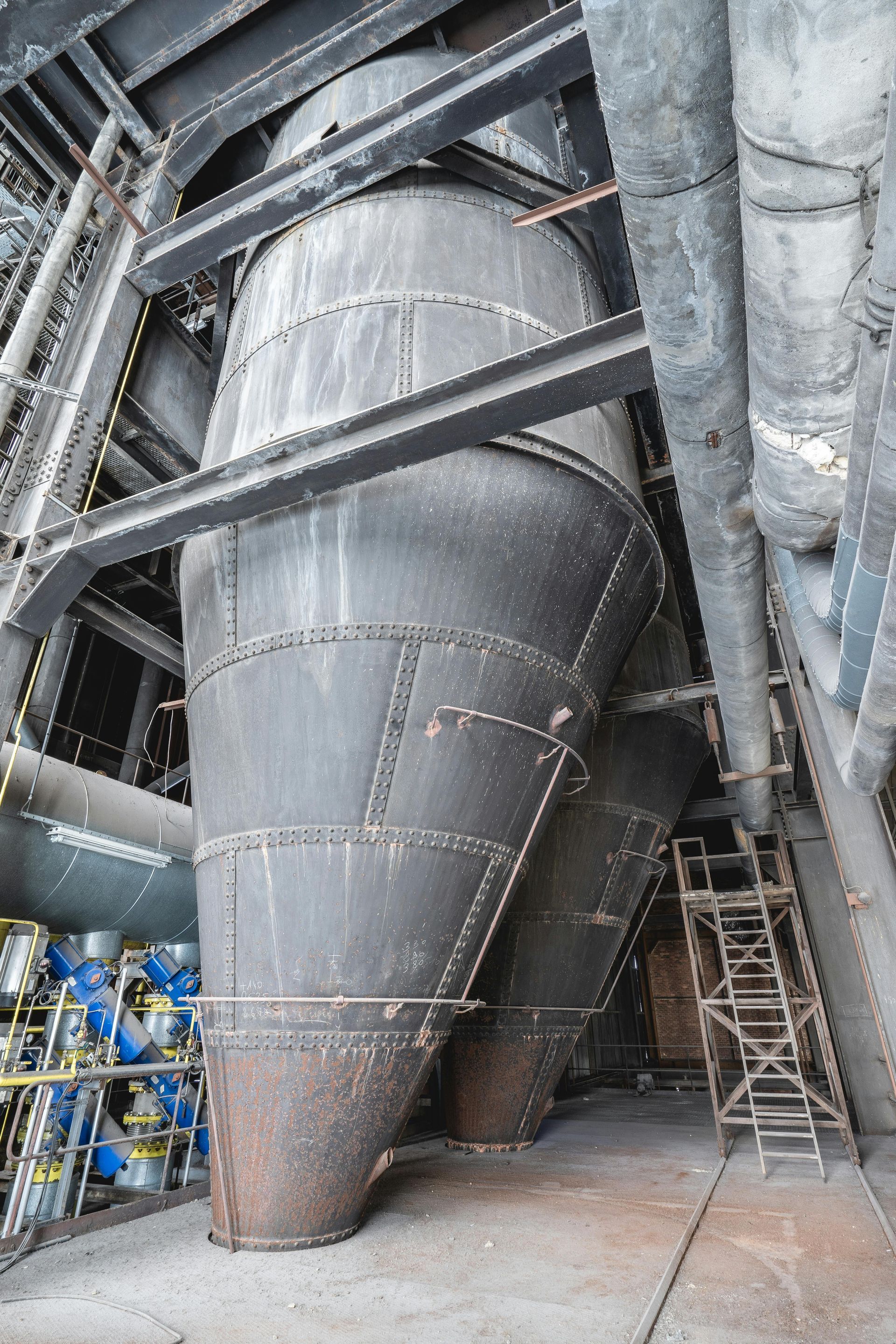 Large, riveted metal industrial hoppers with conical bottoms positioned in a factory setting with support beams.
