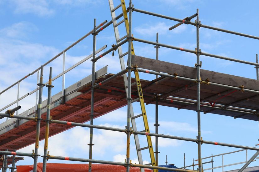 Metal scaffolding structure against a blue sky, featuring wooden planks, safety rails, and a yellow ladder.