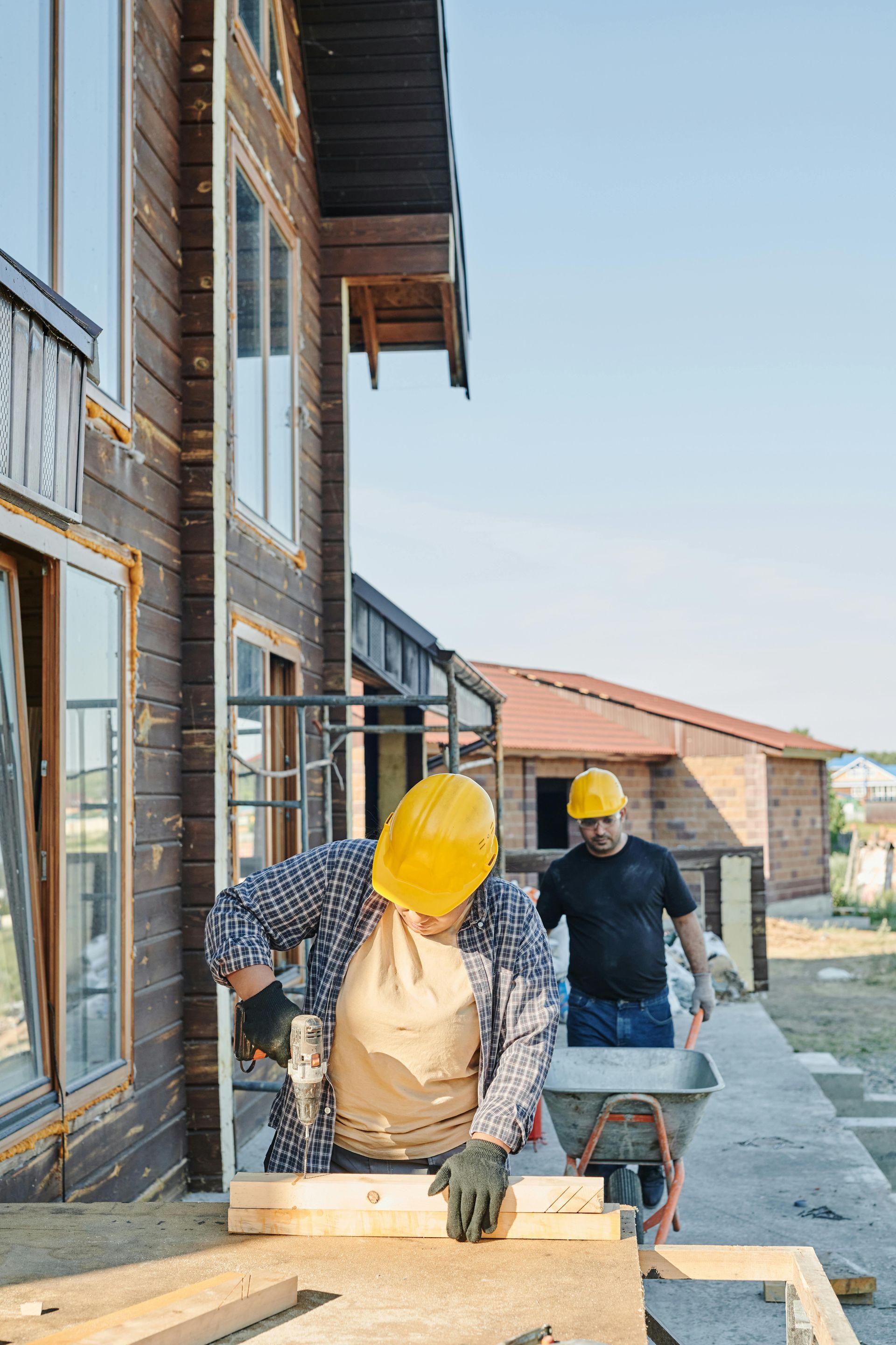 Two construction workers in hard hats work outside a wooden building; one uses a drill on a wooden board.