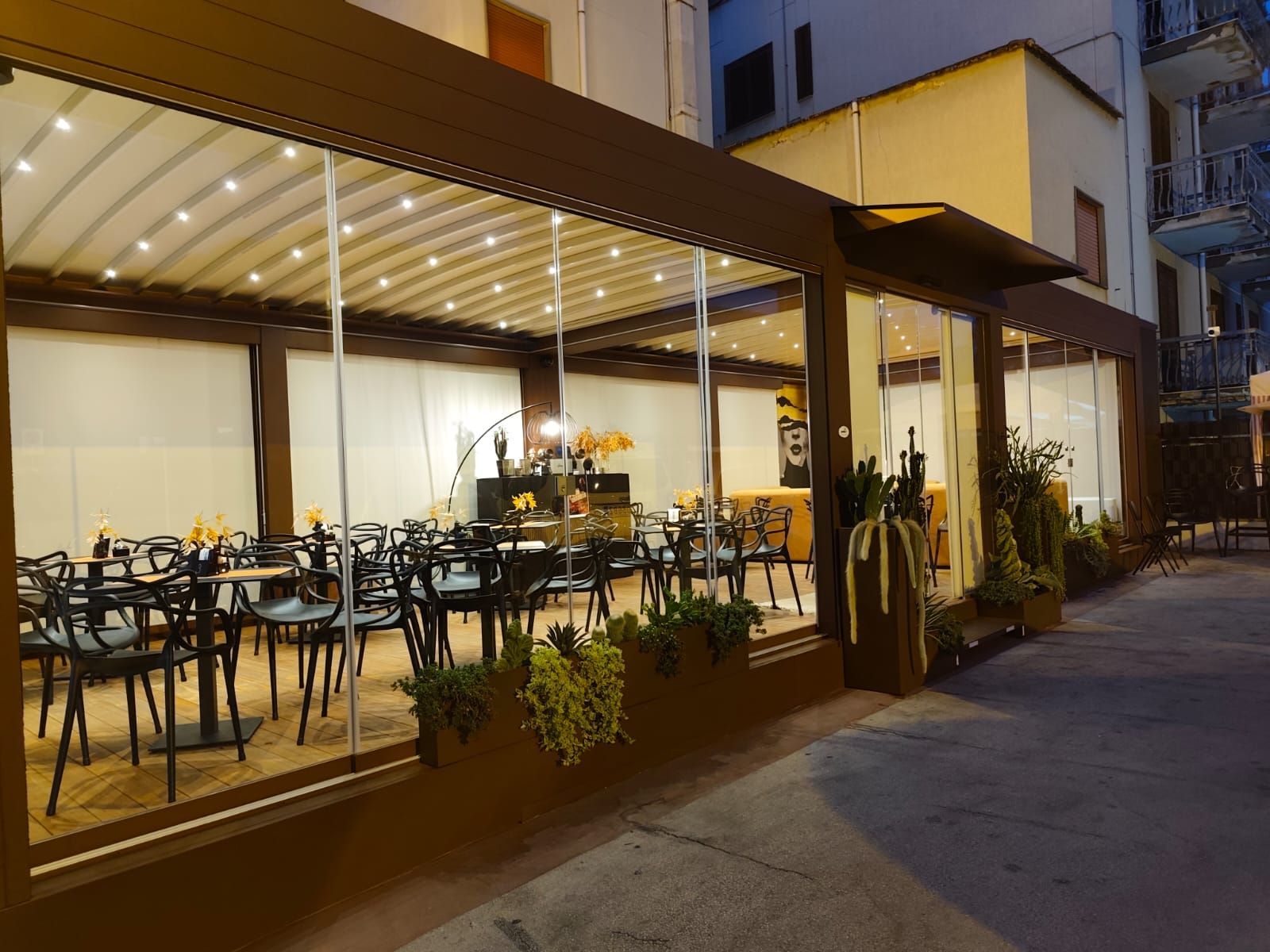 Patio del ristorante con pareti in vetro, tenda da sole marrone, tavoli, sedie e piante. Sera.