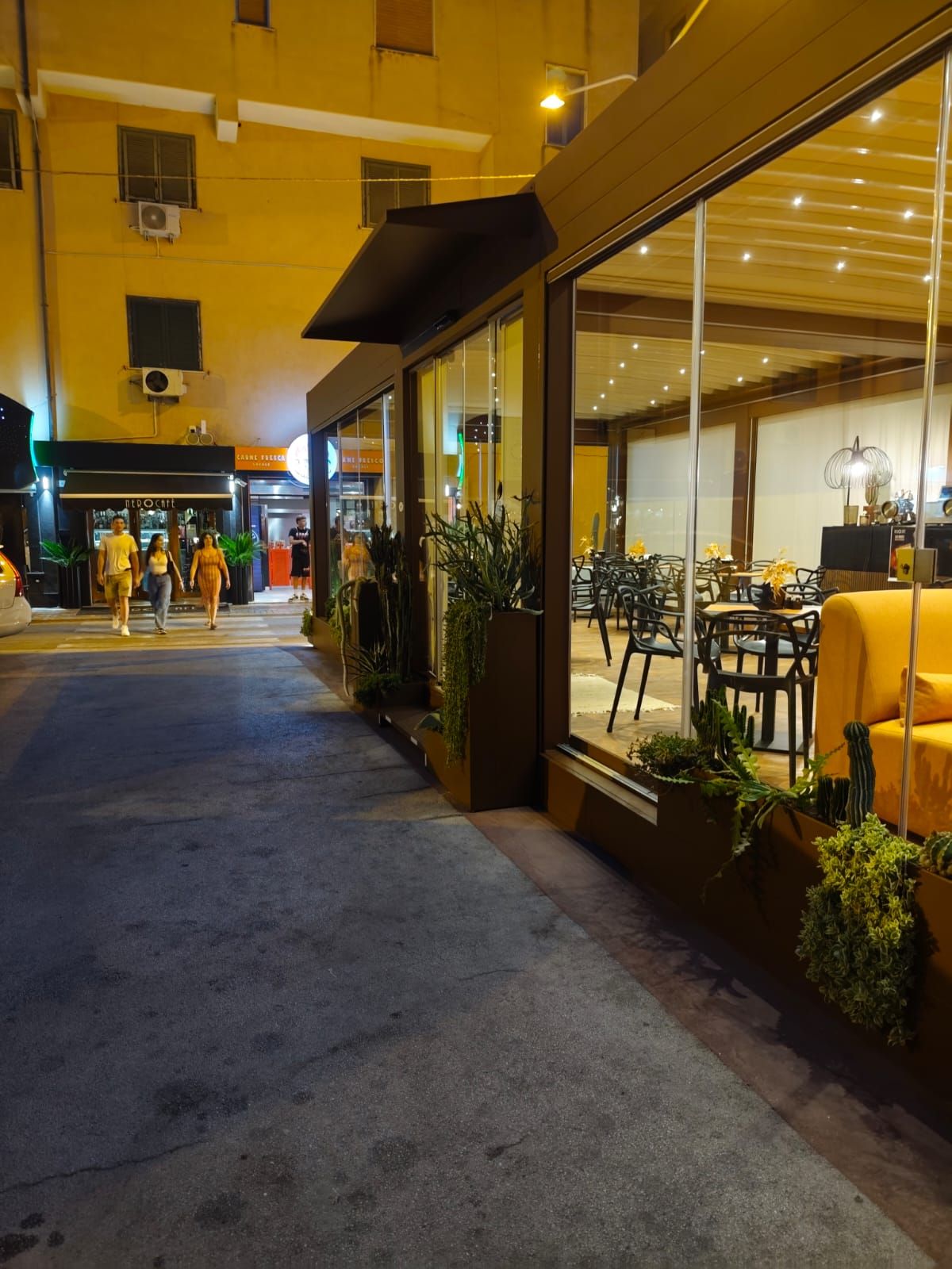 Esterno del ristorante con pareti in vetro, posti a sedere all'aperto e gente che cammina di notte.