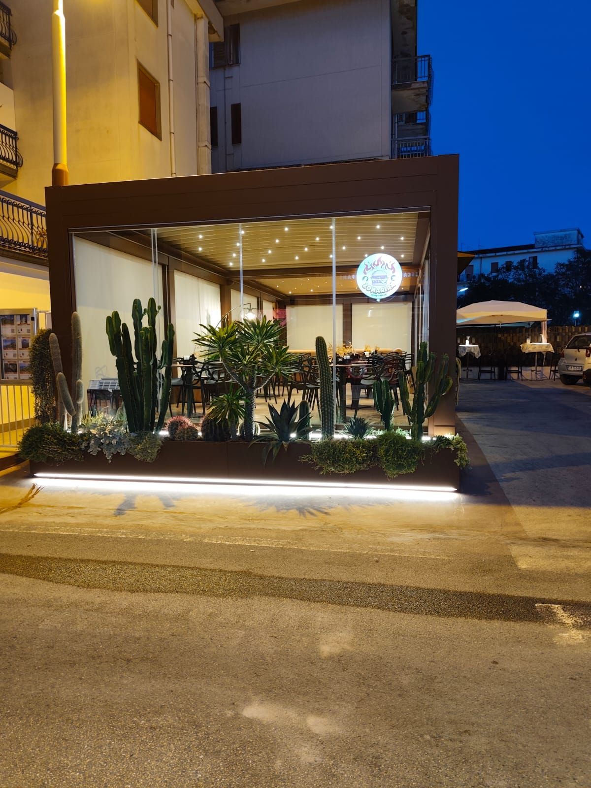 Ristorante con pareti in vetro, cactus e illuminazione notturna dall'alto. Cornice scura, interni bianchi, decorazioni a forma di luna blu.