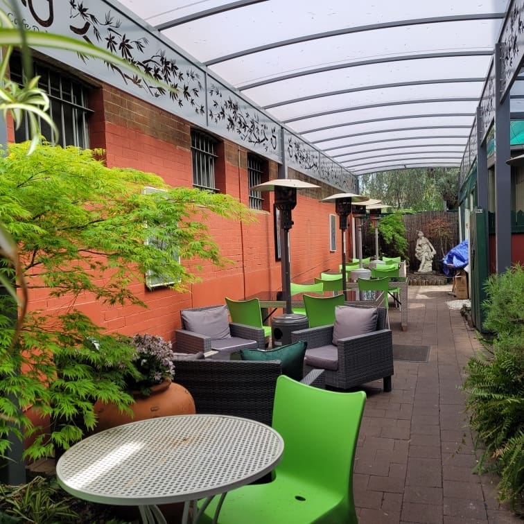 Outdoor cafe with seating, heaters, and canopy. Red brick wall with decorative metalwork. Green and grey furniture.