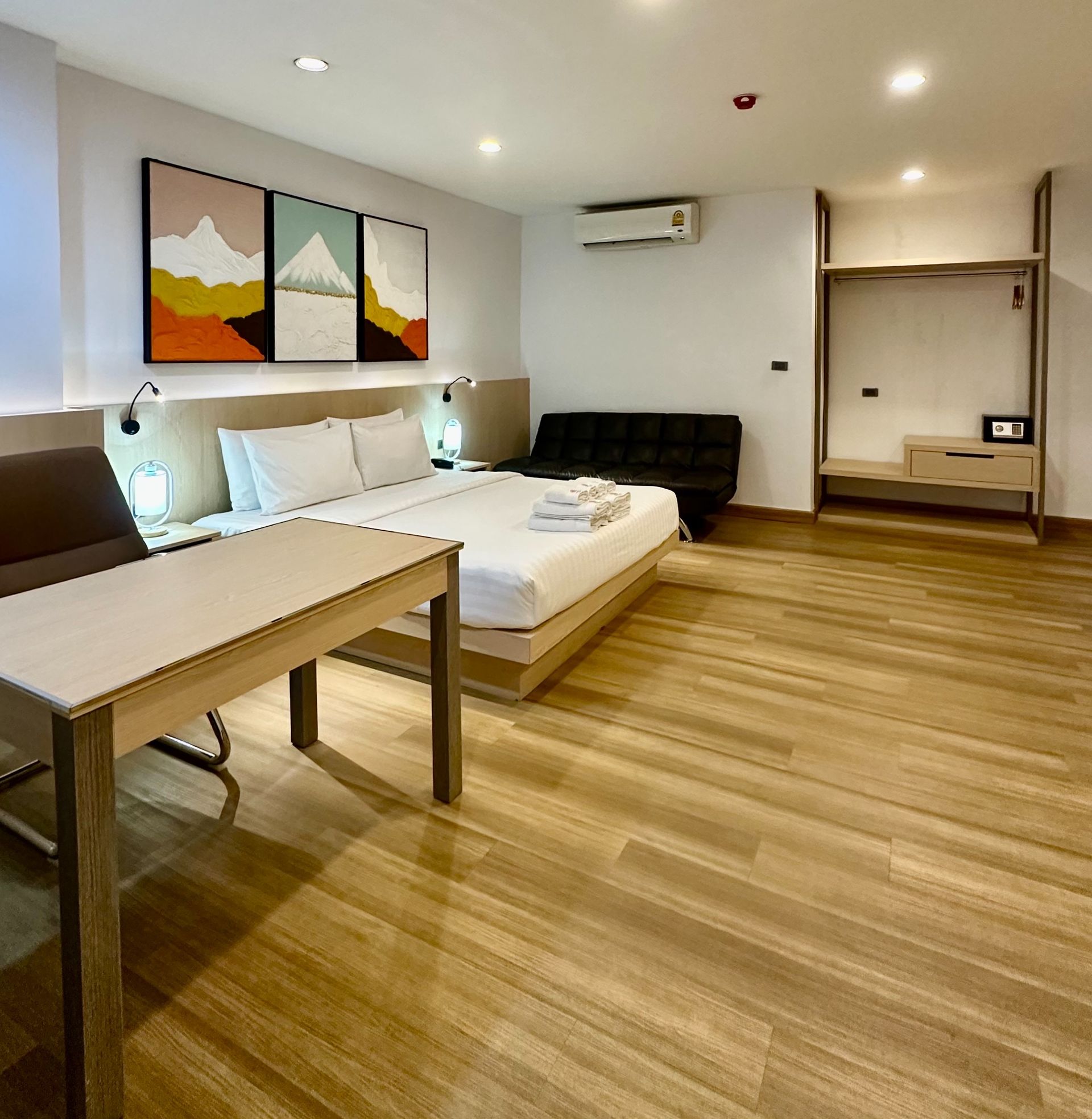Hotel room with bed, desk, sofa, wardrobe, and artwork on the wall. Light wood floors and neutral colors.