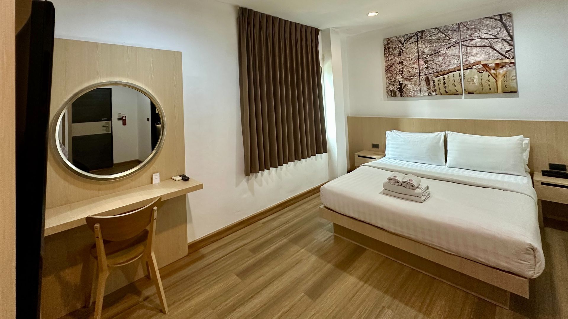 Hotel room with bed, desk, mirror, and artwork. Wooden floors and curtains.