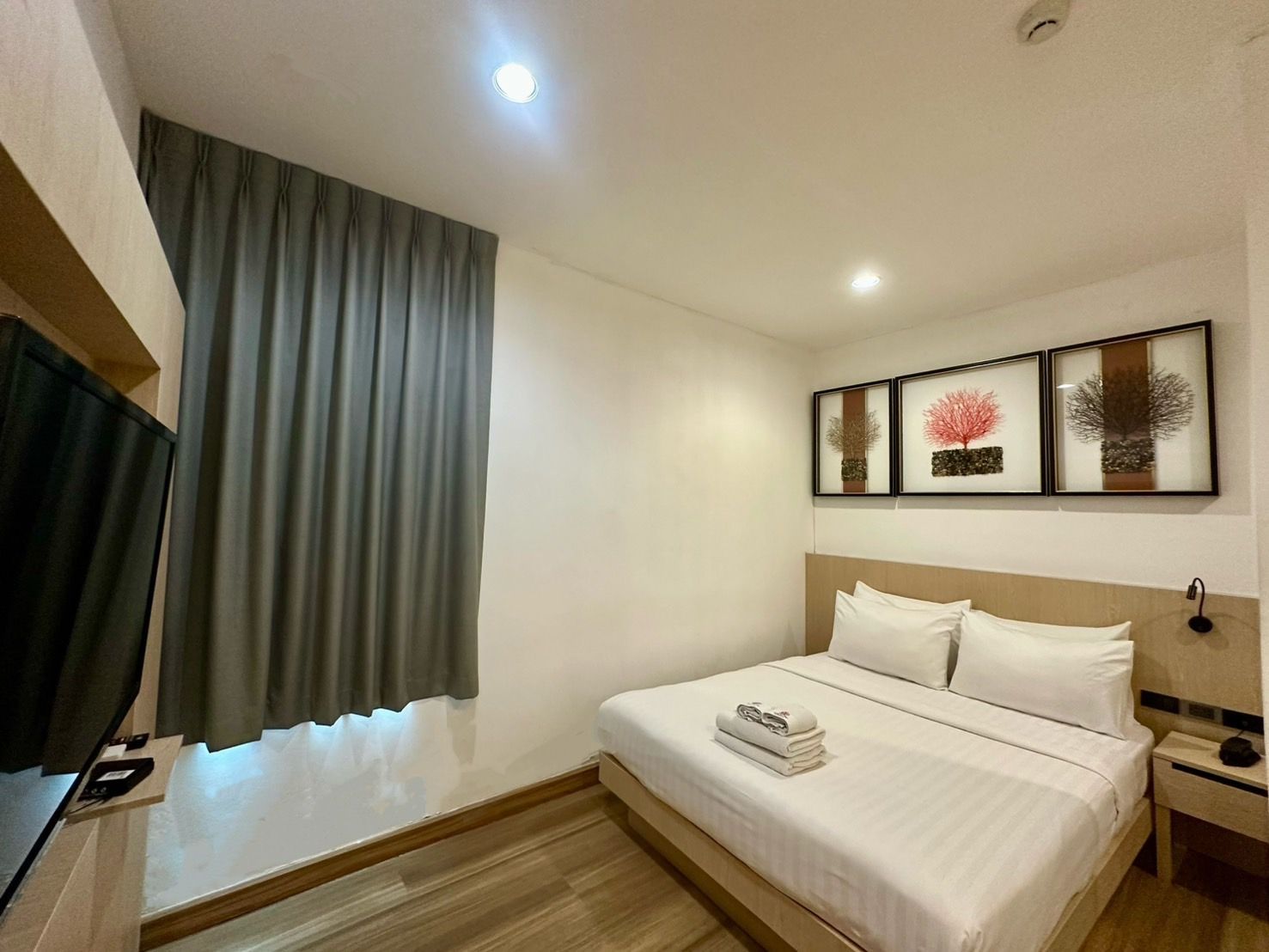 Small hotel room with a bed, TV, and artwork. Grey curtains, white walls, and wood floor.