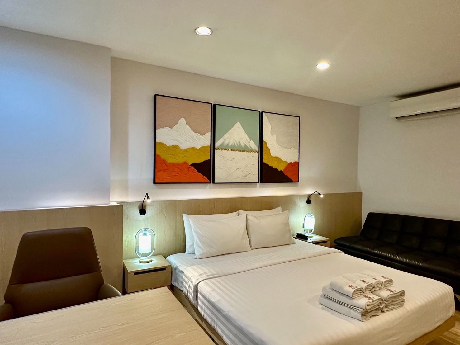 Bedroom with a bed, desk, sofa, and mountain art. Neutral colors, modern style.