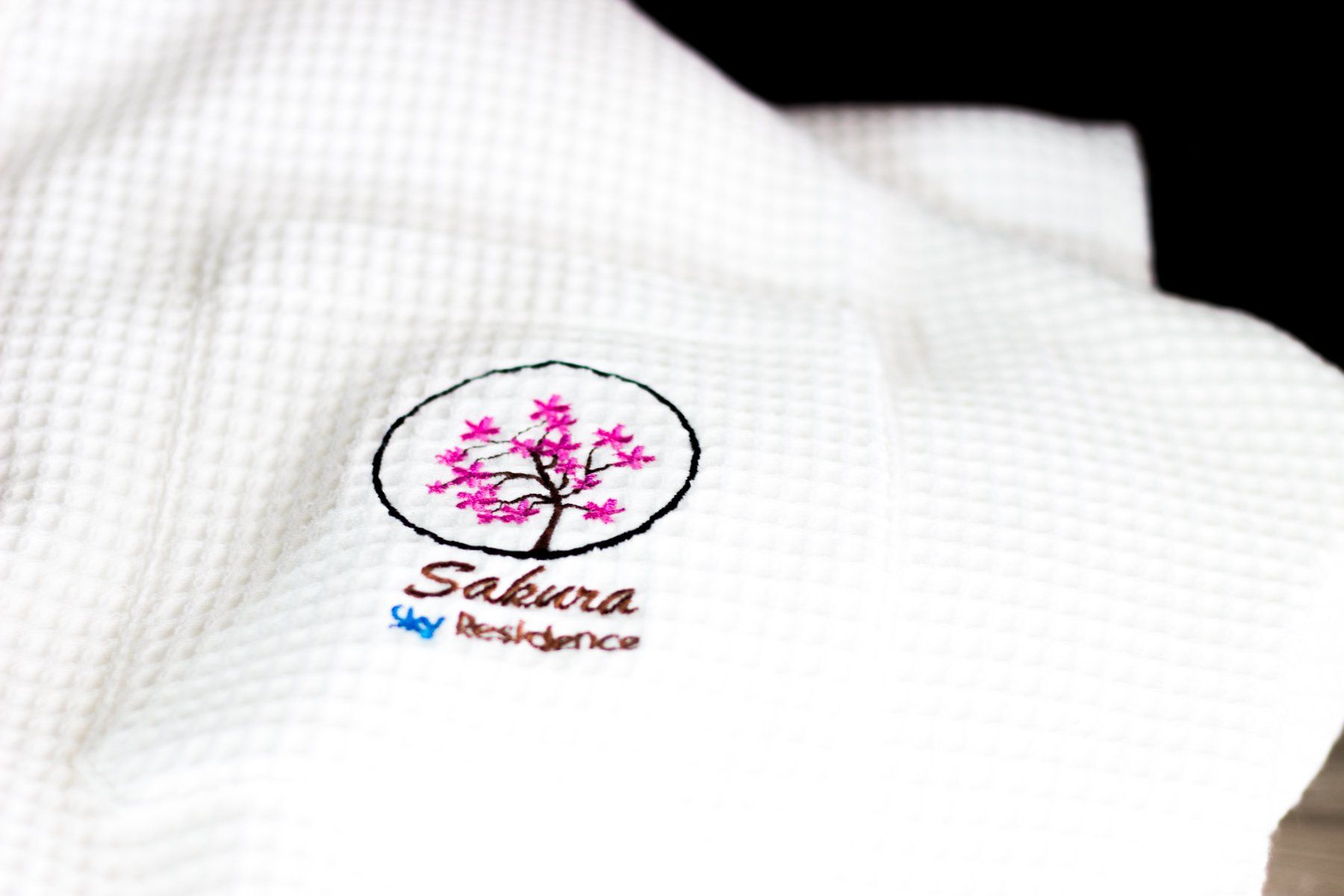 White waffle-textured bathrobe with a Sakura Residence logo embroidered on it: pink cherry blossom in an oval frame.