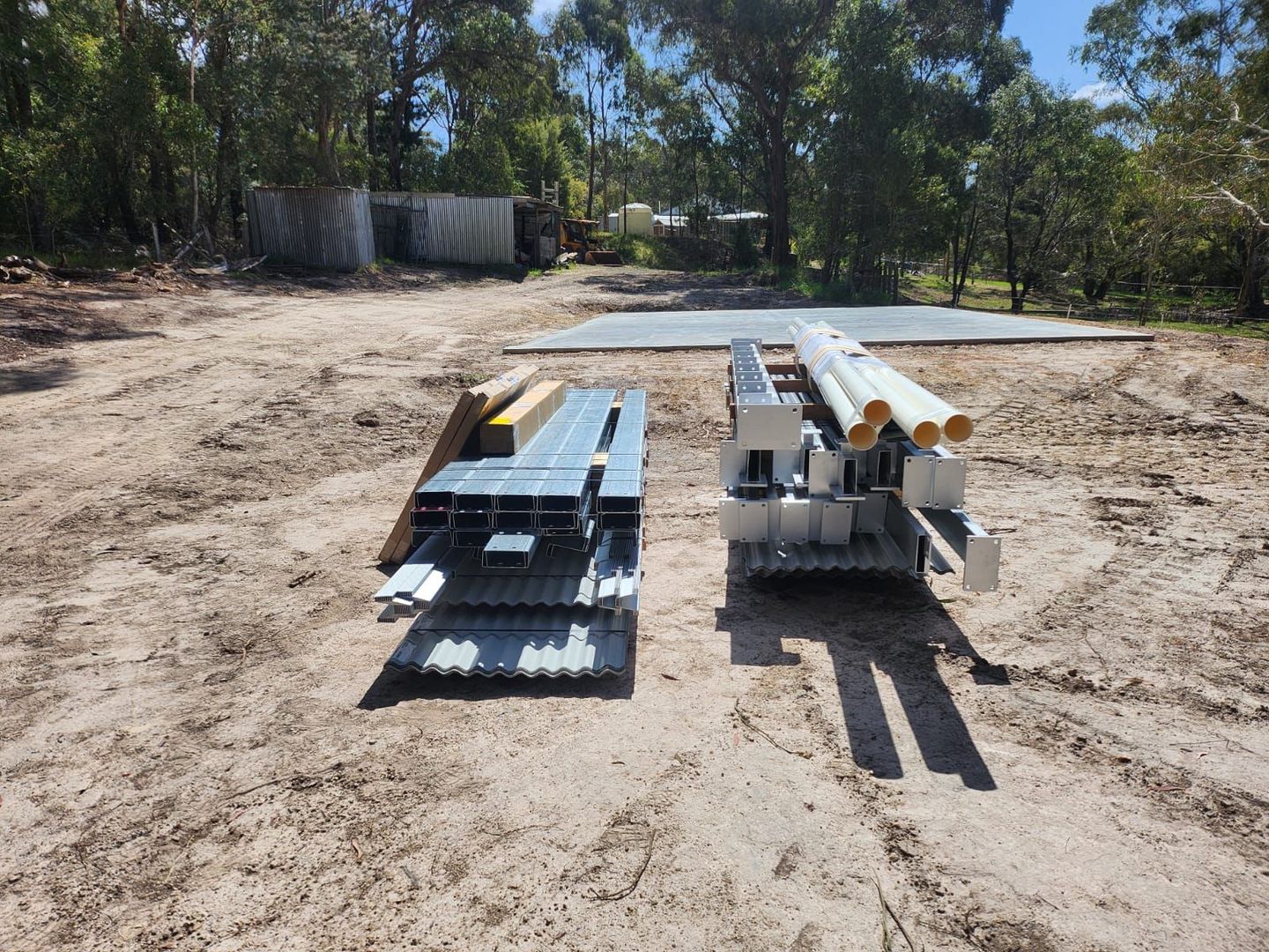 Stacks of metal pipes and panels on pallets in a dirt clearing with trees in the background