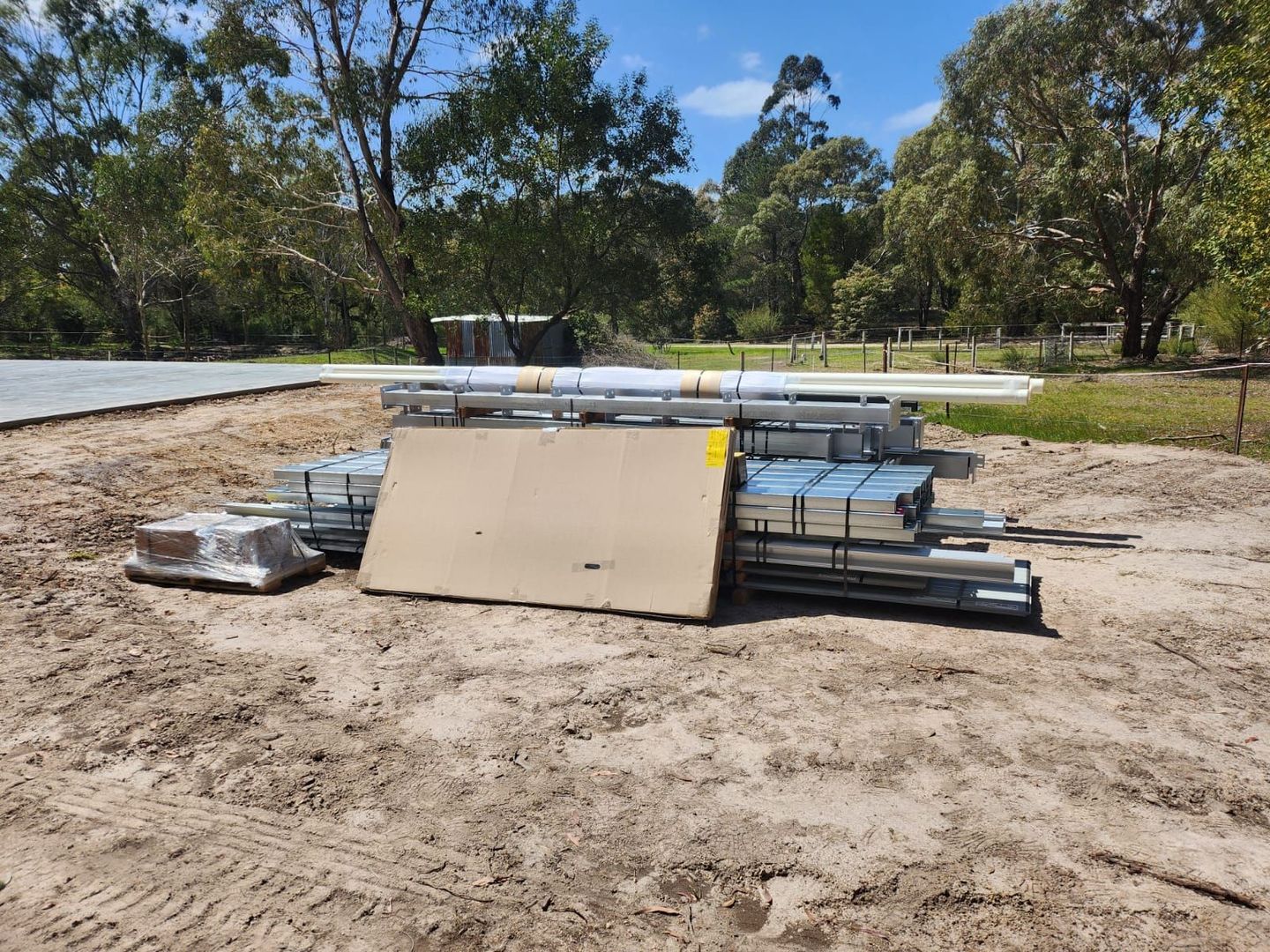 Pallets of construction materials stacked on dirt in a wooded outdoor area