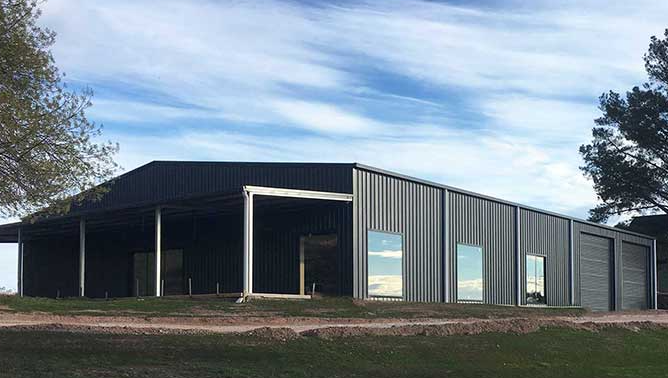 Modern dark metal barn-style building with large windows on a grassy lot under a blue sky